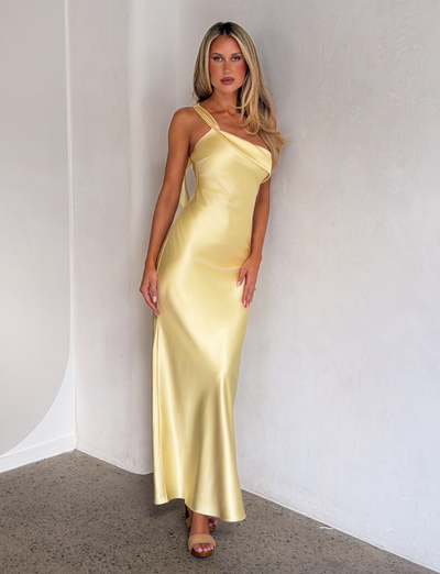A woman with long blonde hair wears the TIGERMIST RAINEY MAXI DRESS in butter yellow and open-toe heels, standing against a light-colored wall on a polished concrete floor.