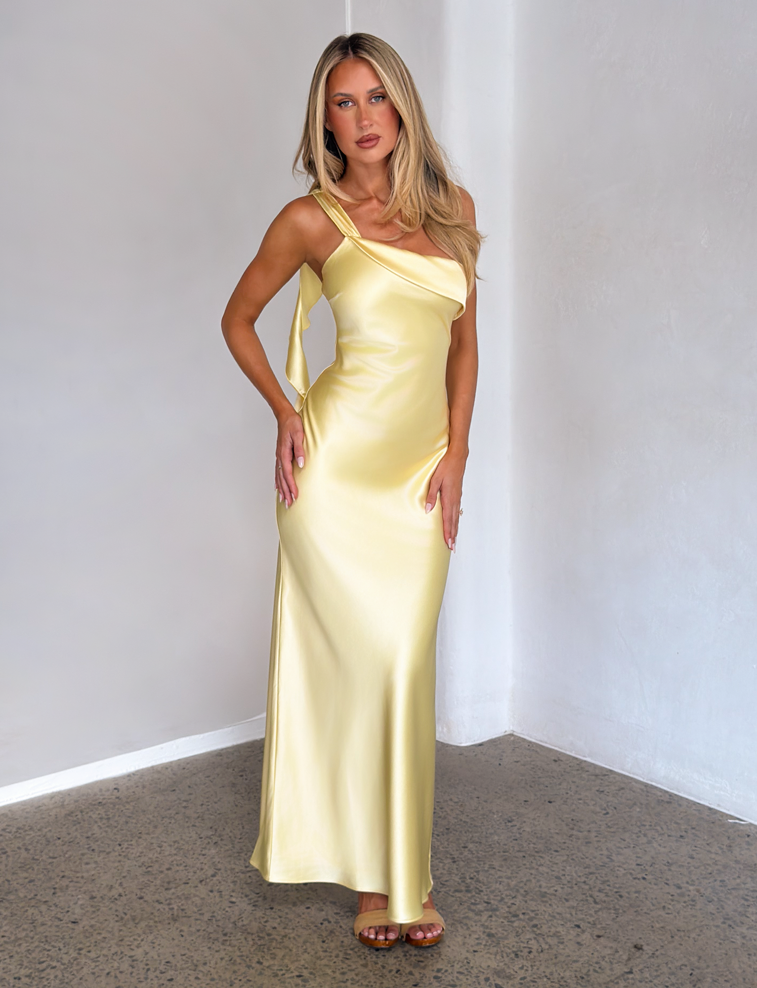 A woman with long blonde hair wears the TIGERMIST RAINEY MAXI DRESS in butter yellow and brown sandals, standing on a polished concrete floor against a light-colored wall.