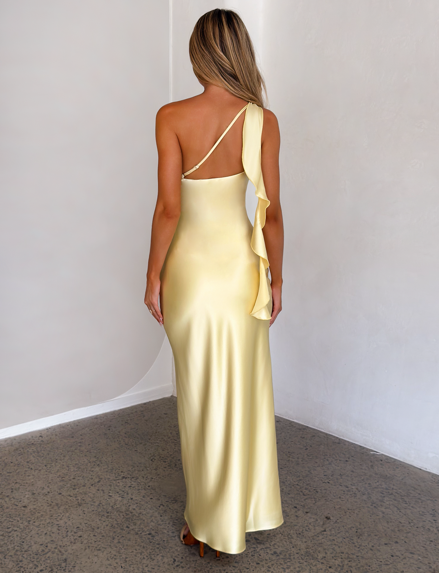 A woman stands indoors with her back to the camera wearing the TIGERMIST RAINEY MAXI DRESS in butter yellow, a one-shoulder satin dress with elegant waterfall ruffle details, against plain light walls on a speckled floor.