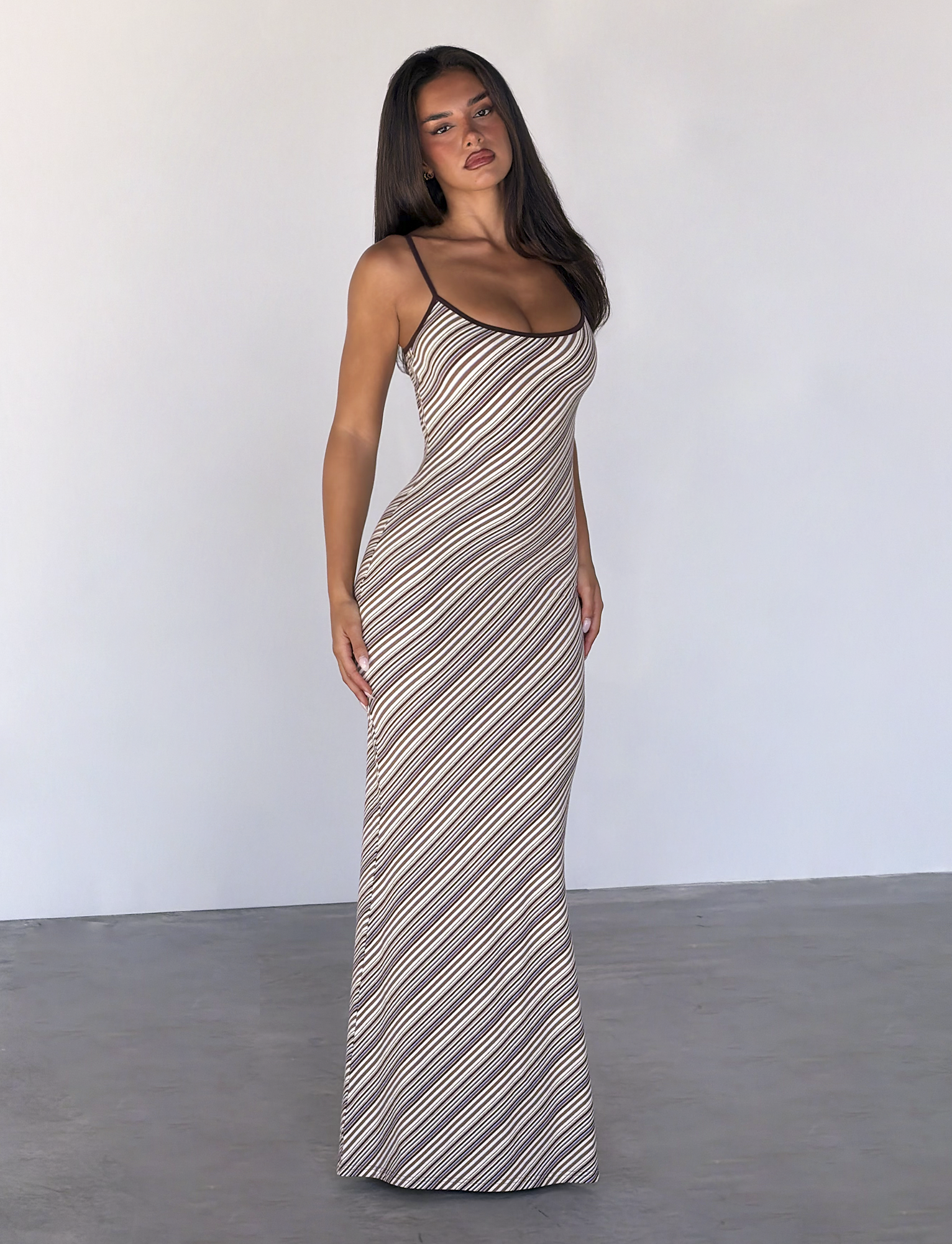 A woman poses confidently indoors against a plain light wall, wearing the TIGERMIST SLOANE MAXI DRESS in tan and choc stripe.