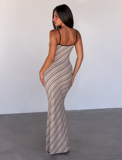 A woman with long dark hair stands on a smooth concrete floor, facing away and looking over her shoulder, wearing the TIGERMIST SLOANE MAXI DRESS in tan and choc stripe against a plain light-colored background.