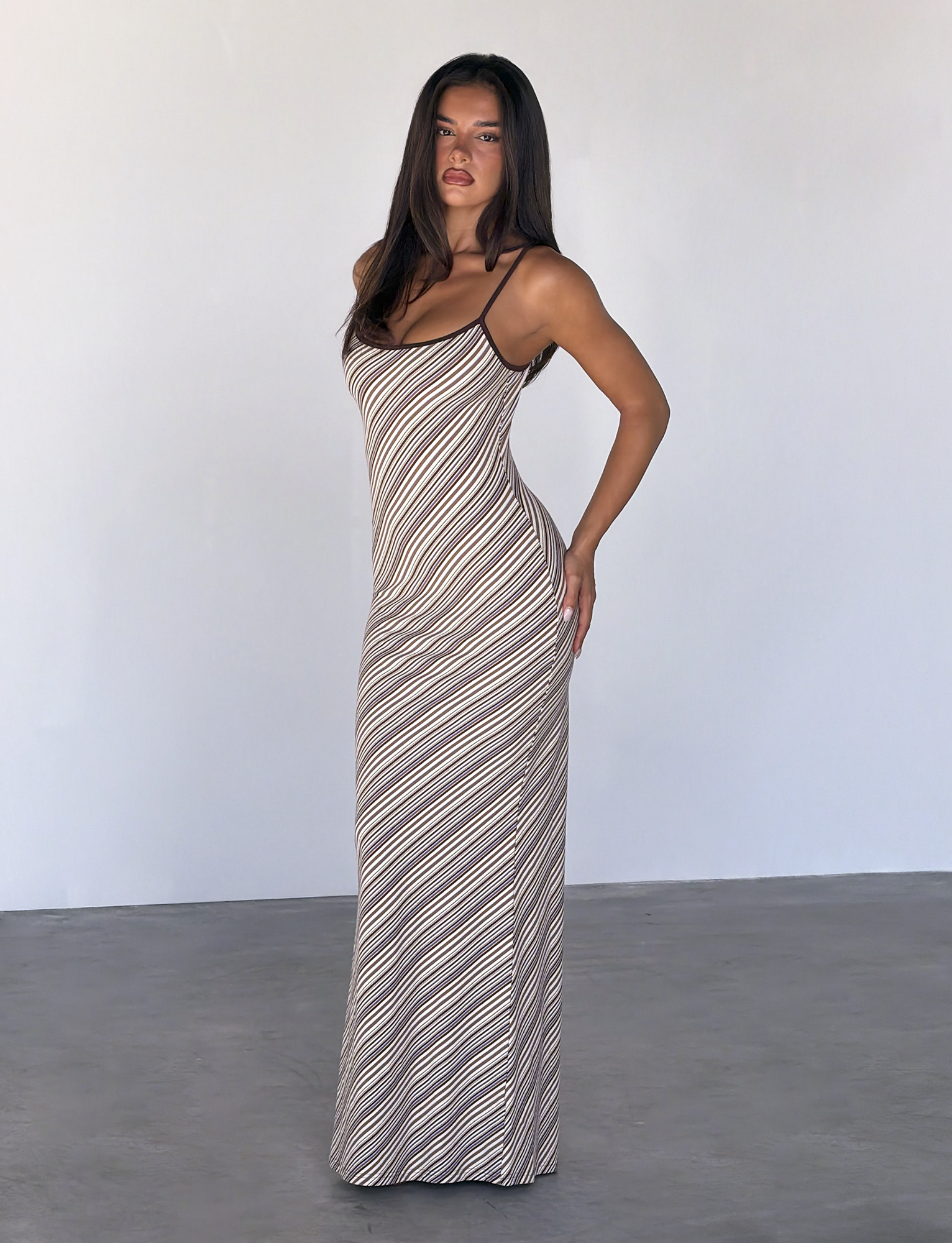 A woman with long dark hair stands indoors against a plain light wall, wearing the TIGERMIST SLOANE MAXI DRESS in Tan and Choc Stripe. She poses confidently with one hand on her hip and looks at the camera.