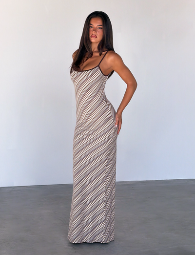 A woman with long dark hair stands indoors against a plain light wall, wearing the TIGERMIST SLOANE MAXI DRESS in Tan and Choc Stripe. She poses confidently with one hand on her hip and looks at the camera.