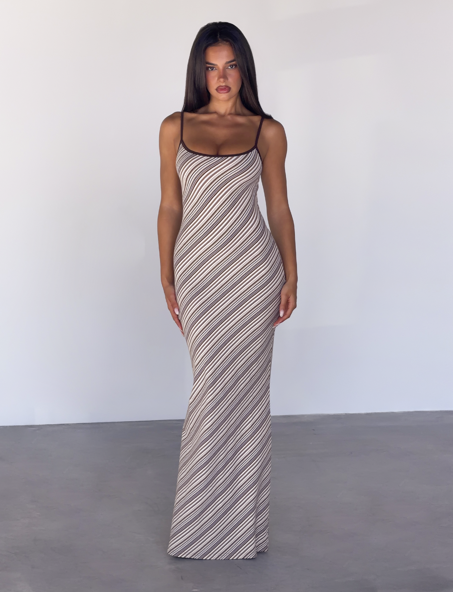 A woman with long dark hair stands facing forward in a plain, light-colored room, wearing the TIGERMIST SLOANE MAXI DRESS in tan and chocolate stripe.