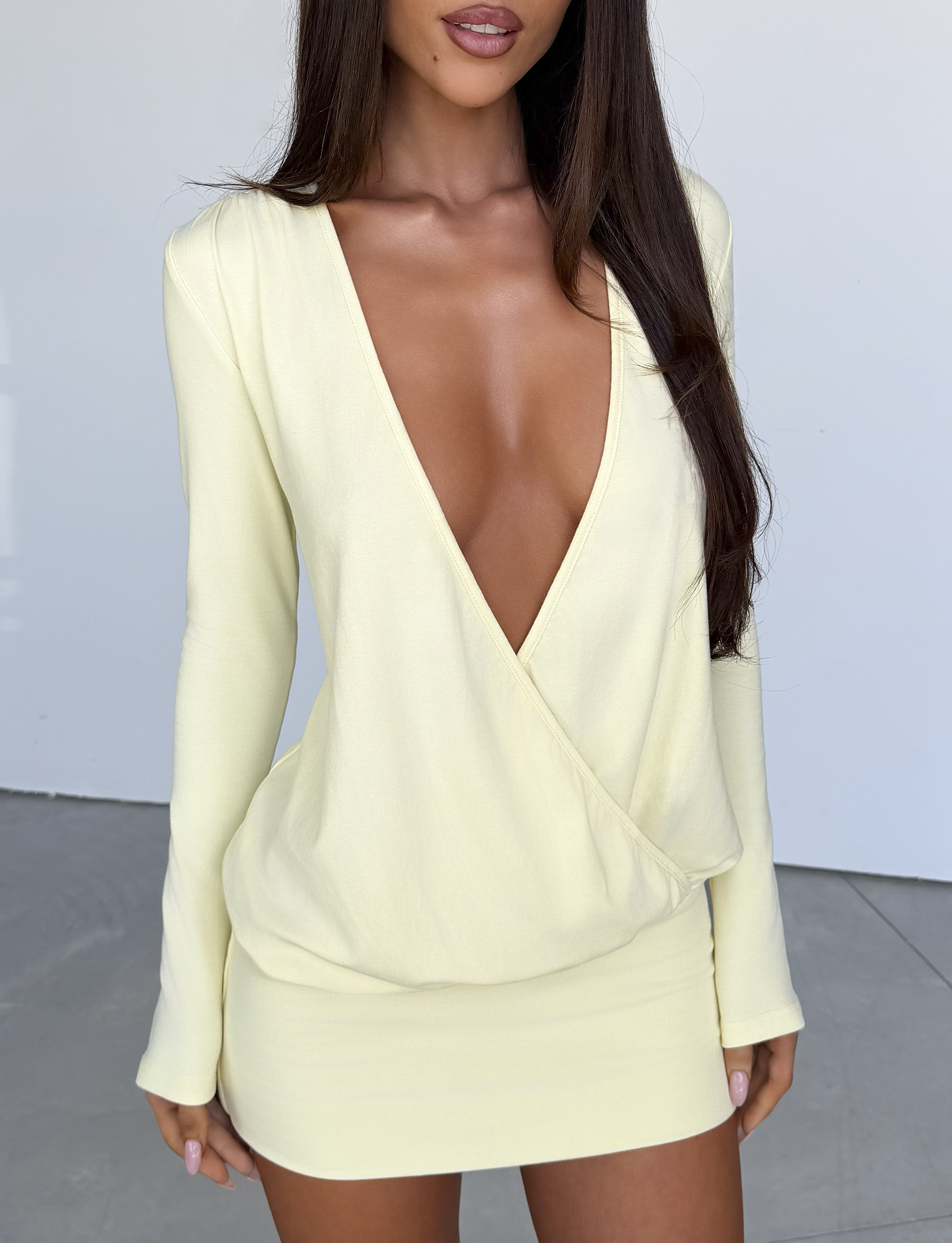 A woman wearing the TIGERMIST FOXLEY MINI DRESS in Butter Cream, featuring long sleeves and a deep V-neckline, stands against a plain, light-colored background. Her face is partially visible, and her long dark hair is down.