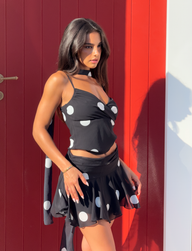 A woman with long dark hair, wearing a black crop top and the TIGERMIST AYDIN MINI SKIRT in black, stands in sunlight by a red door, gazing to the side with a serious expression.