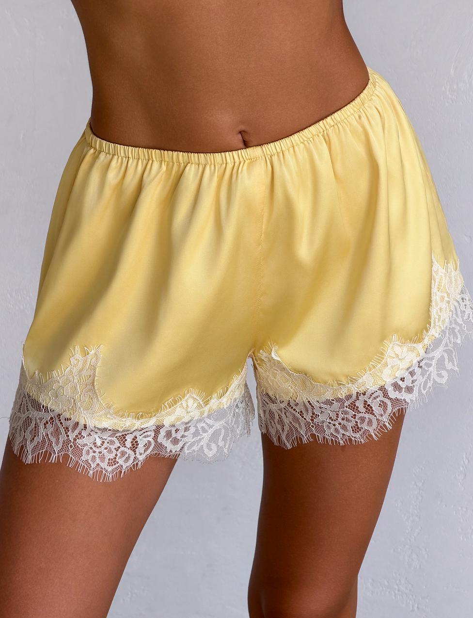 A person wears the TIGERMIST SOLSTICE SHORT in yellow, featuring white lace trim along the hem, and stands against a light-colored background.