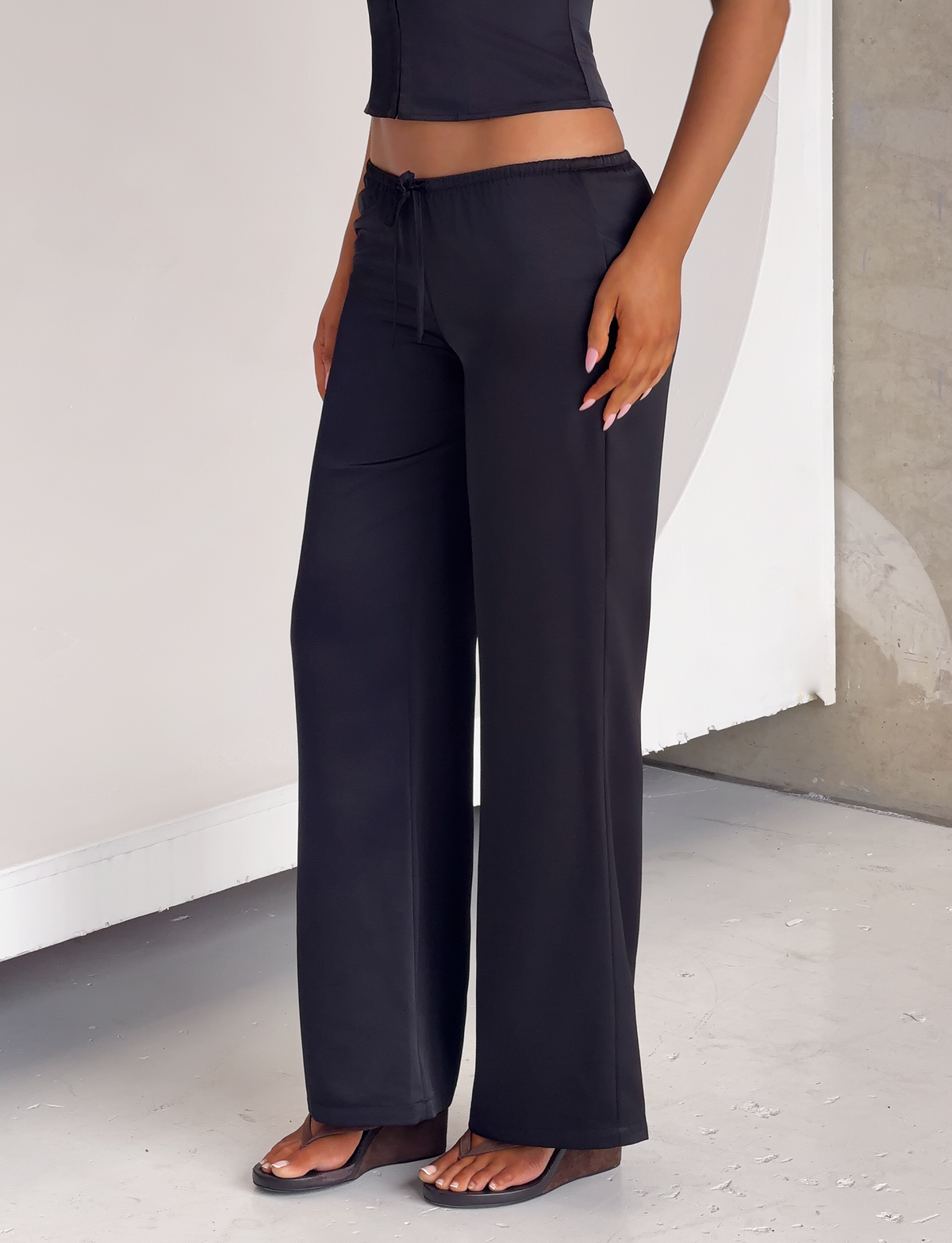 MAURA PANT - BLACK trousers in black satin with a wide-leg cut and adjustable drawstring waist, worn with brown platform sandals.