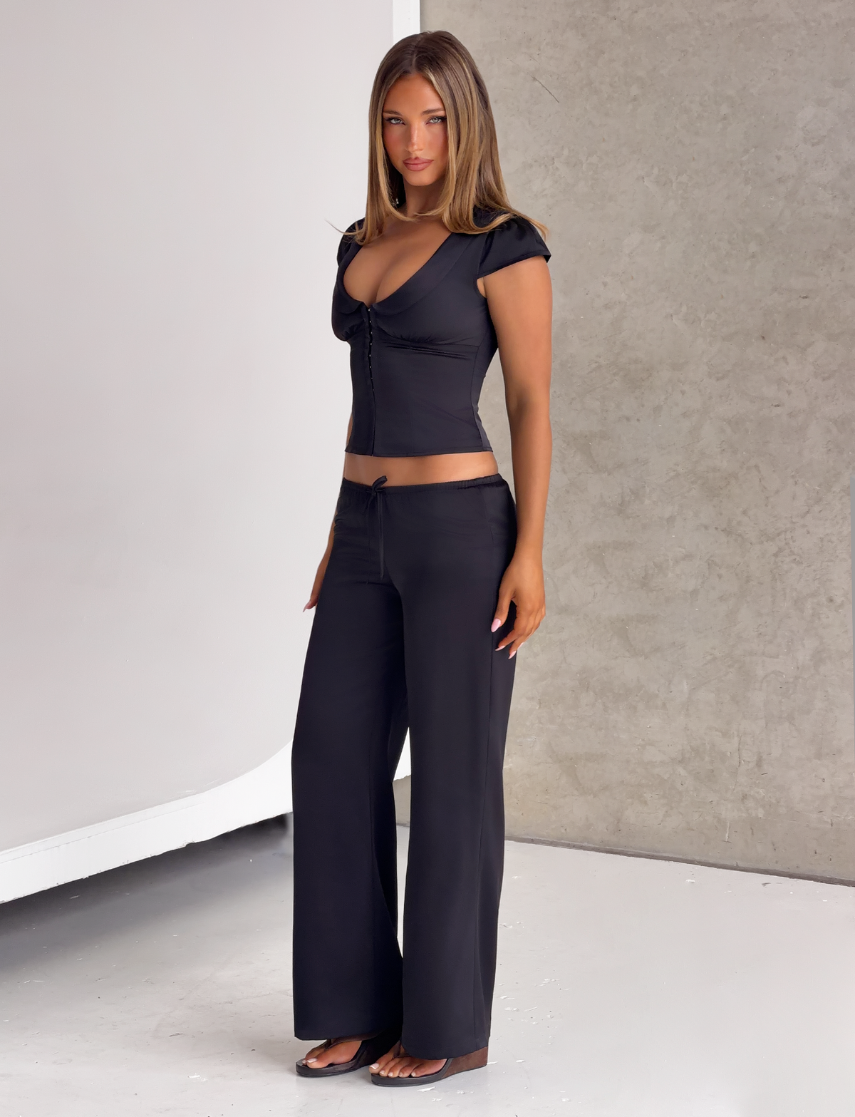 A woman wearing a black short-sleeved top with a front-button detail and matching black MAURA PANT - BLACK wide-leg satin pants.