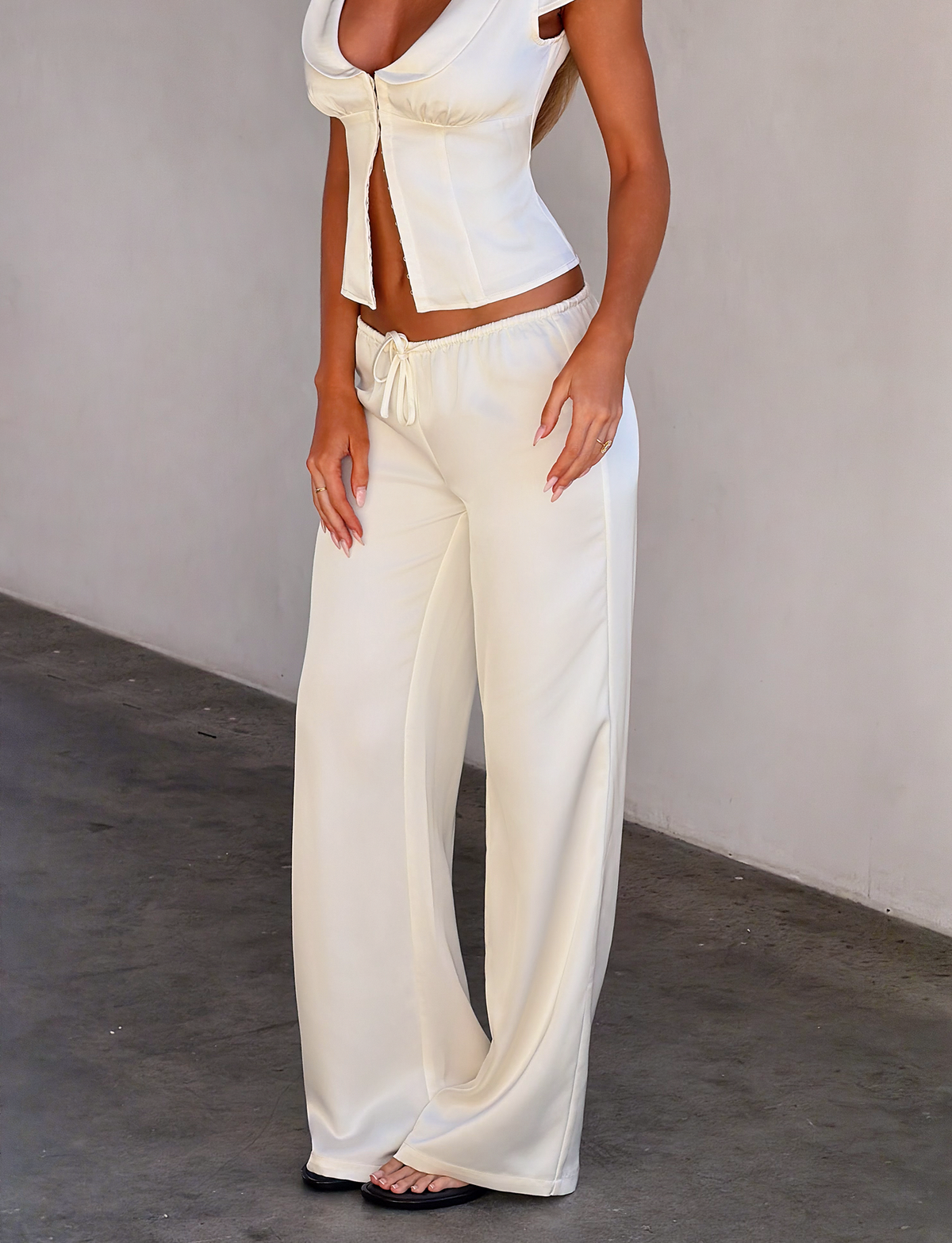 A woman stands against a plain wall wearing TIGERMIST's MAURA PANT in white—a drapey, satin, wide-legged style with an adjustable waist—paired with a stylish cropped top and open-toe sandals. Her face is not visible in the image.