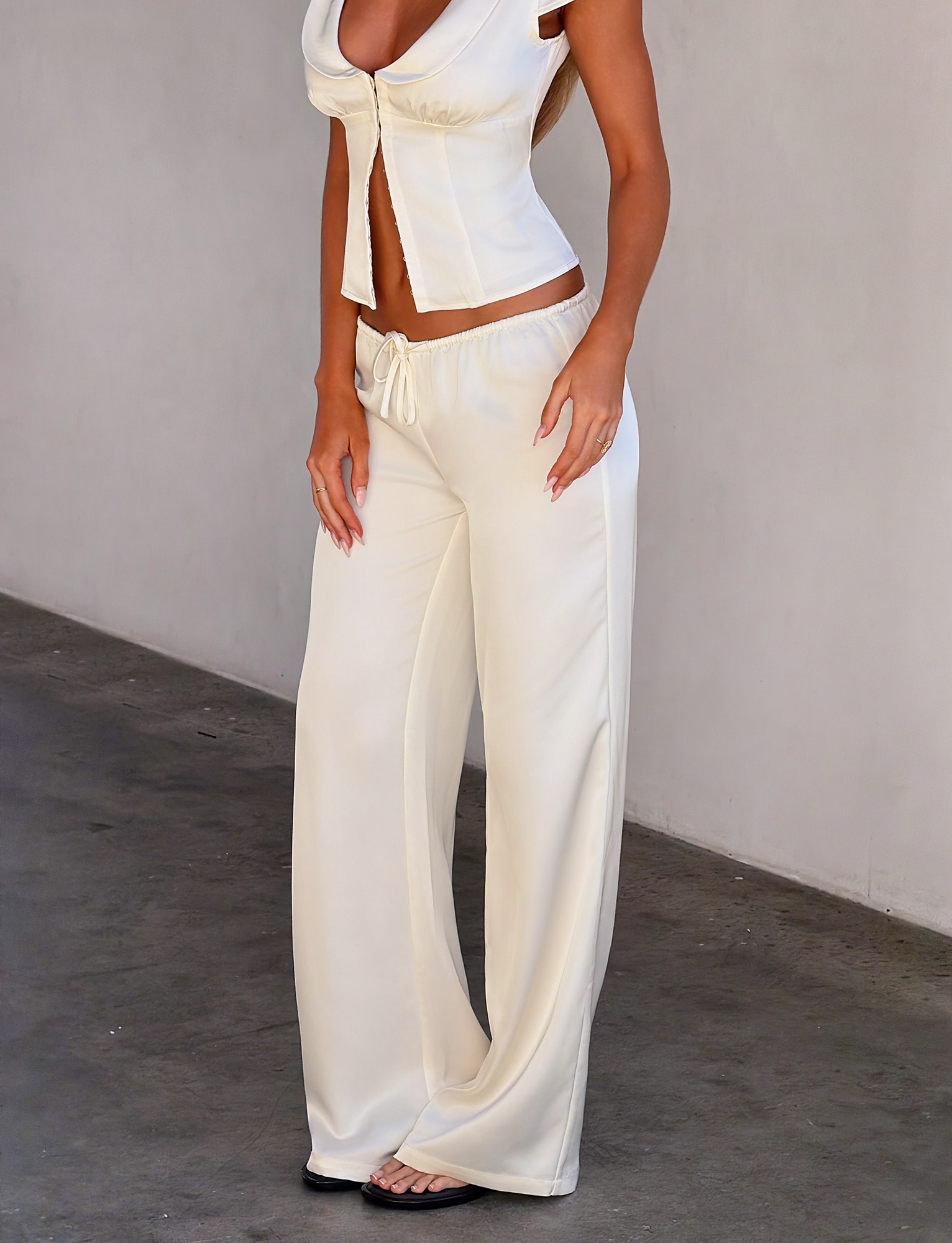 A woman stands against a plain wall wearing TIGERMIST's MAURA PANT in white—a drapey, satin, wide-legged style with an adjustable waist—paired with a stylish cropped top and open-toe sandals. Her face is not visible in the image.