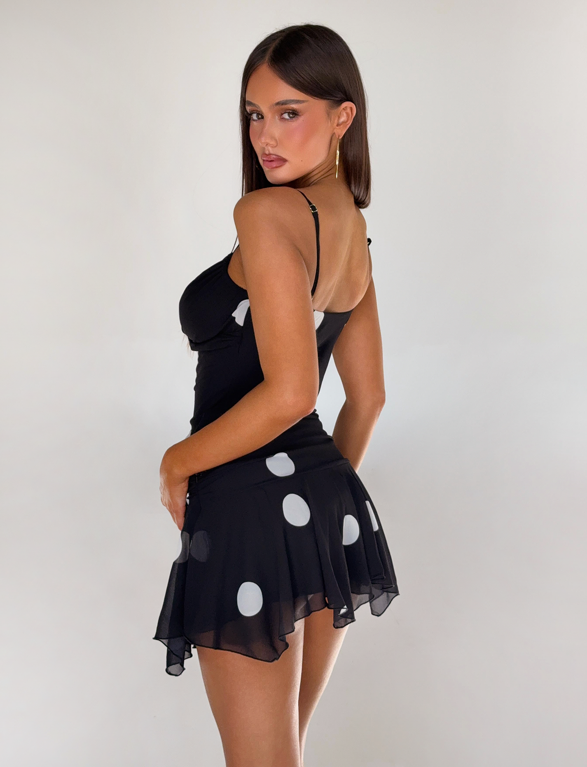 A woman with long brown hair poses against a plain white background wearing the TIGERMIST AYDIN MINI SKIRT in black, a sleeveless style with a polka dot pattern featuring large white dots.
