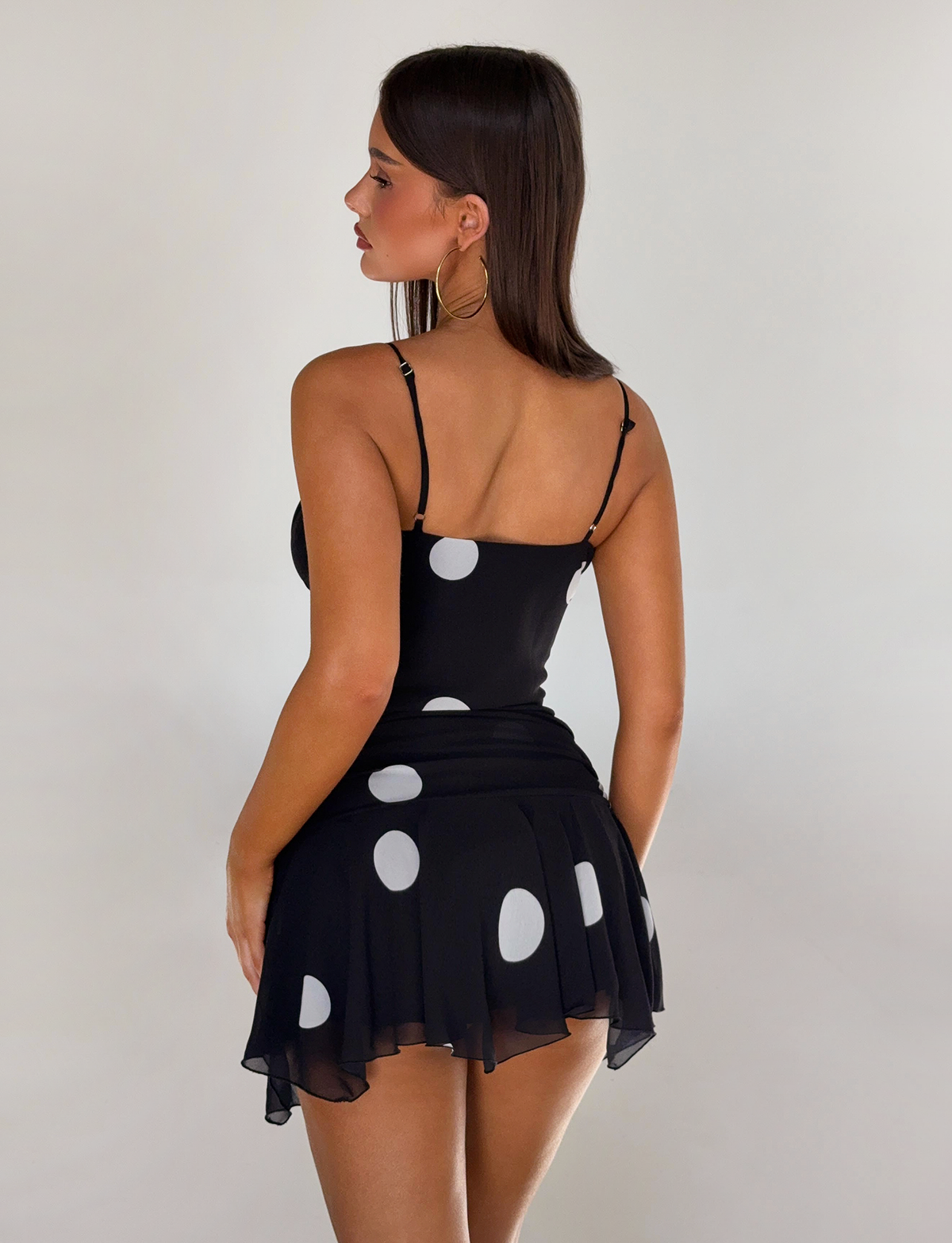 A woman with straight dark hair models the TIGERMIST AYDIN MINI SKIRT - BLACK, a flowy chiffon mini skirt with large white polka dots. She is standing against a plain light background, facing away and slightly to the side.