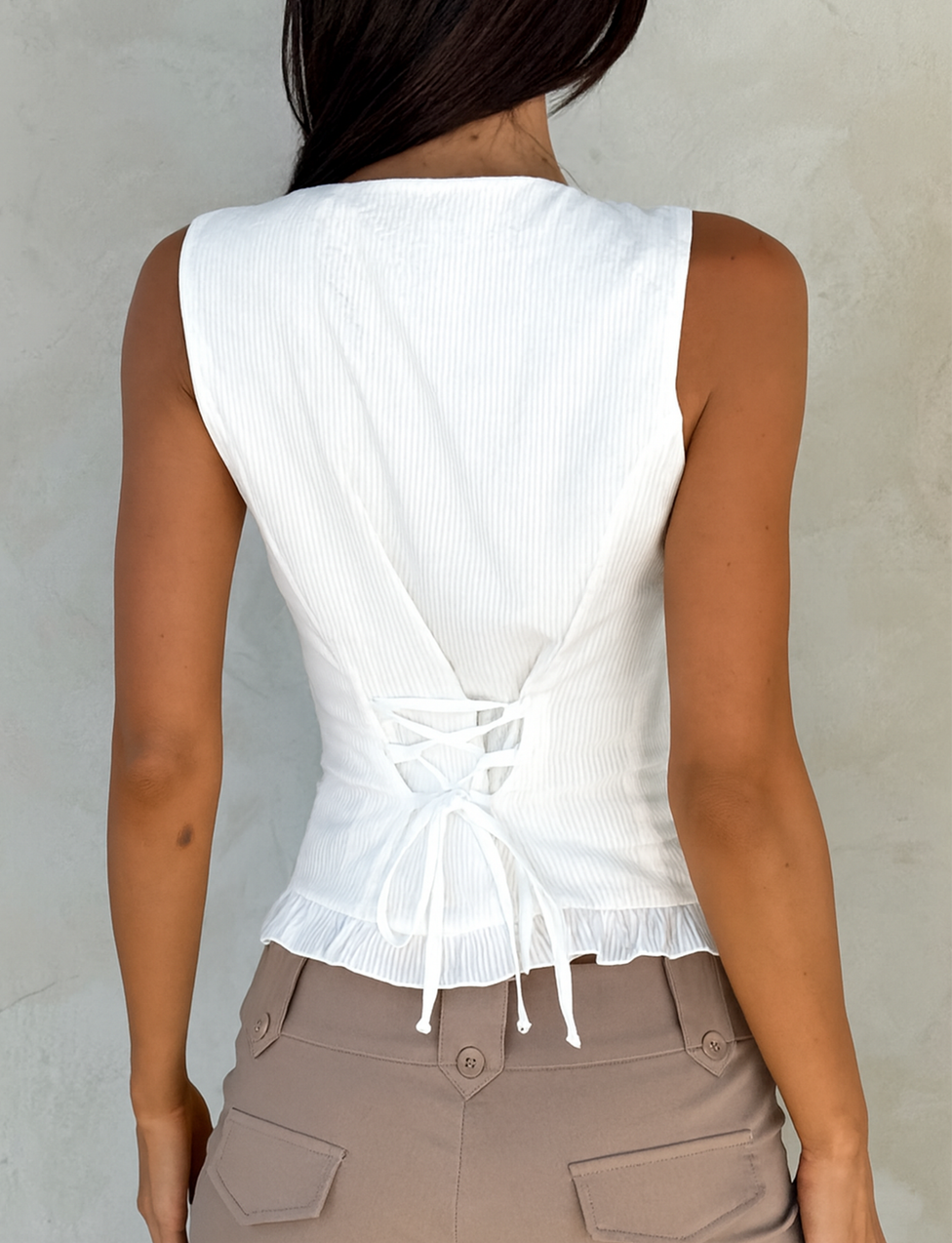 A woman is seen from the back wearing the TIGERMIST ANIYA KEYHOLE TOP in white, featuring a sleeveless cut and lace-up waist, paired with beige high-waisted pants against a light gray wall.