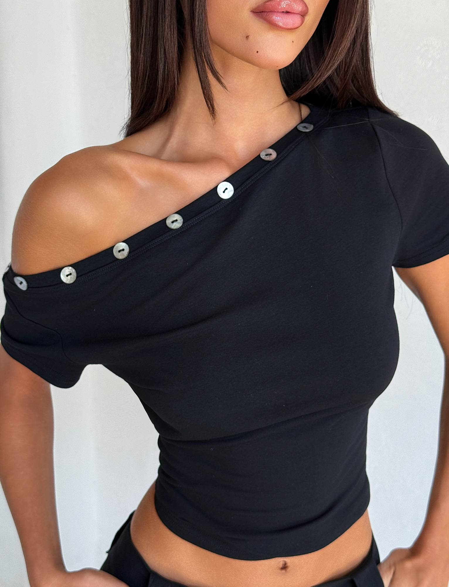 A woman models the TIGERMIST MARITA TOP in black, featuring an off-the-shoulder design and button accents along the asymmetric neckline. Her straight brown hair stands out against a plain white background.