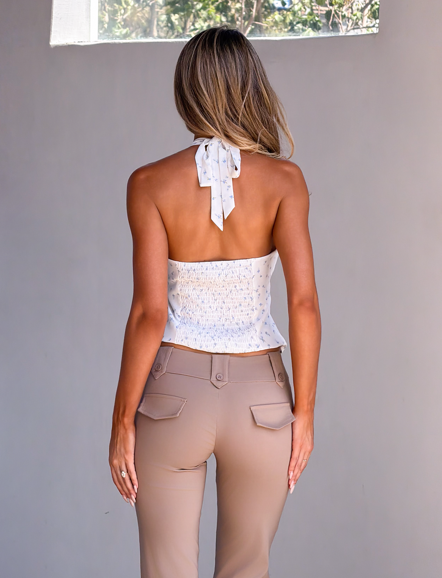 A woman with long blonde hair faces away, wearing the TIGERMIST CALI TOP - DITSY FLORAL with a bow back and high-waisted beige pants, standing against a plain wall beneath a rectangular window.
