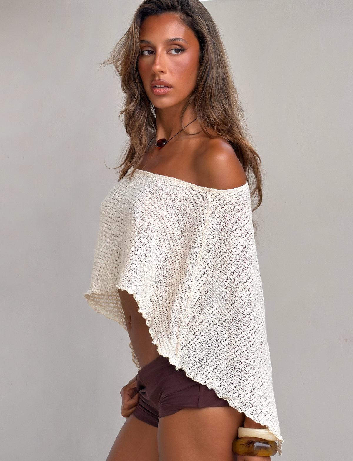 A woman with long hair poses sideways against a plain light background, wearing the TIGERMIST FAIRLEE PONCHO in cream and dark brown shorts.