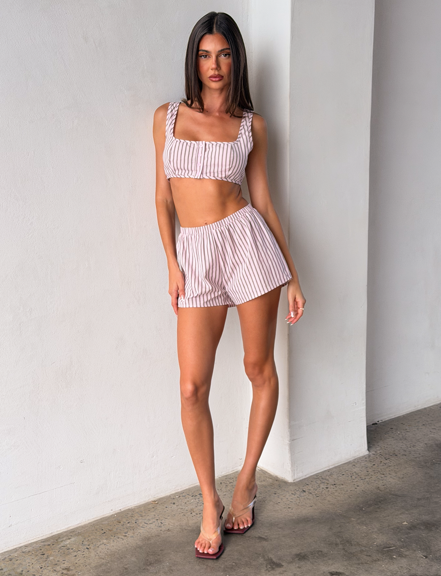 A woman stands indoors against a white wall, wearing the TIGERMIST EZRA TOP - PINK STRIPE and matching shorts with clear heeled sandals.