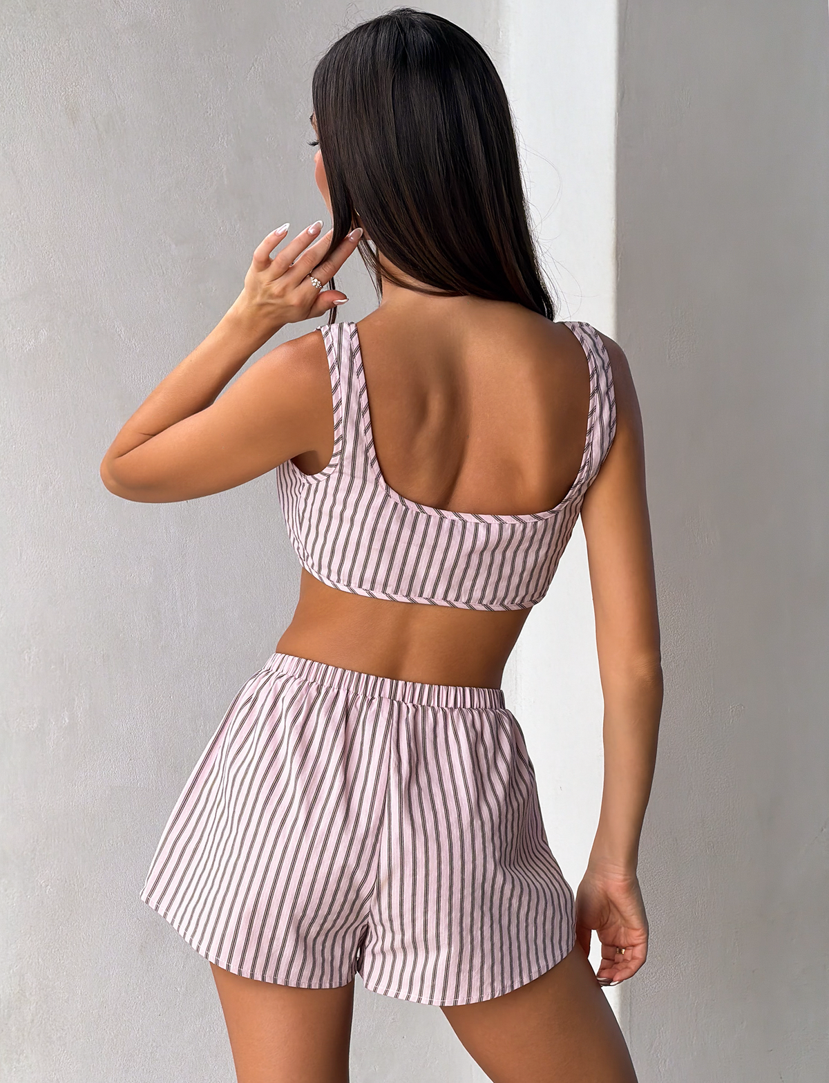 A woman with long brown hair faces a white wall, wearing the TIGERMIST EZRA TOP - PINK STRIPE and matching shorts. Her back is to the camera, and her left hand is raised near her head.