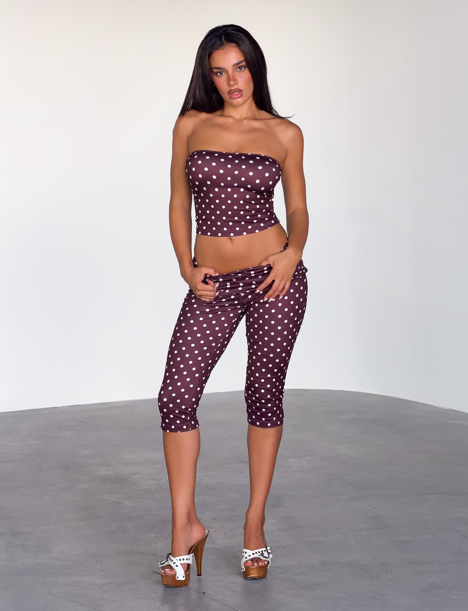 In a minimal room, a woman with long sleek dark hair and a confident look models the TIGERMIST VALI POLKA DOT TOP - CHOCOLATE PINK DOT, paired with matching capri pants and high-heeled sandals.