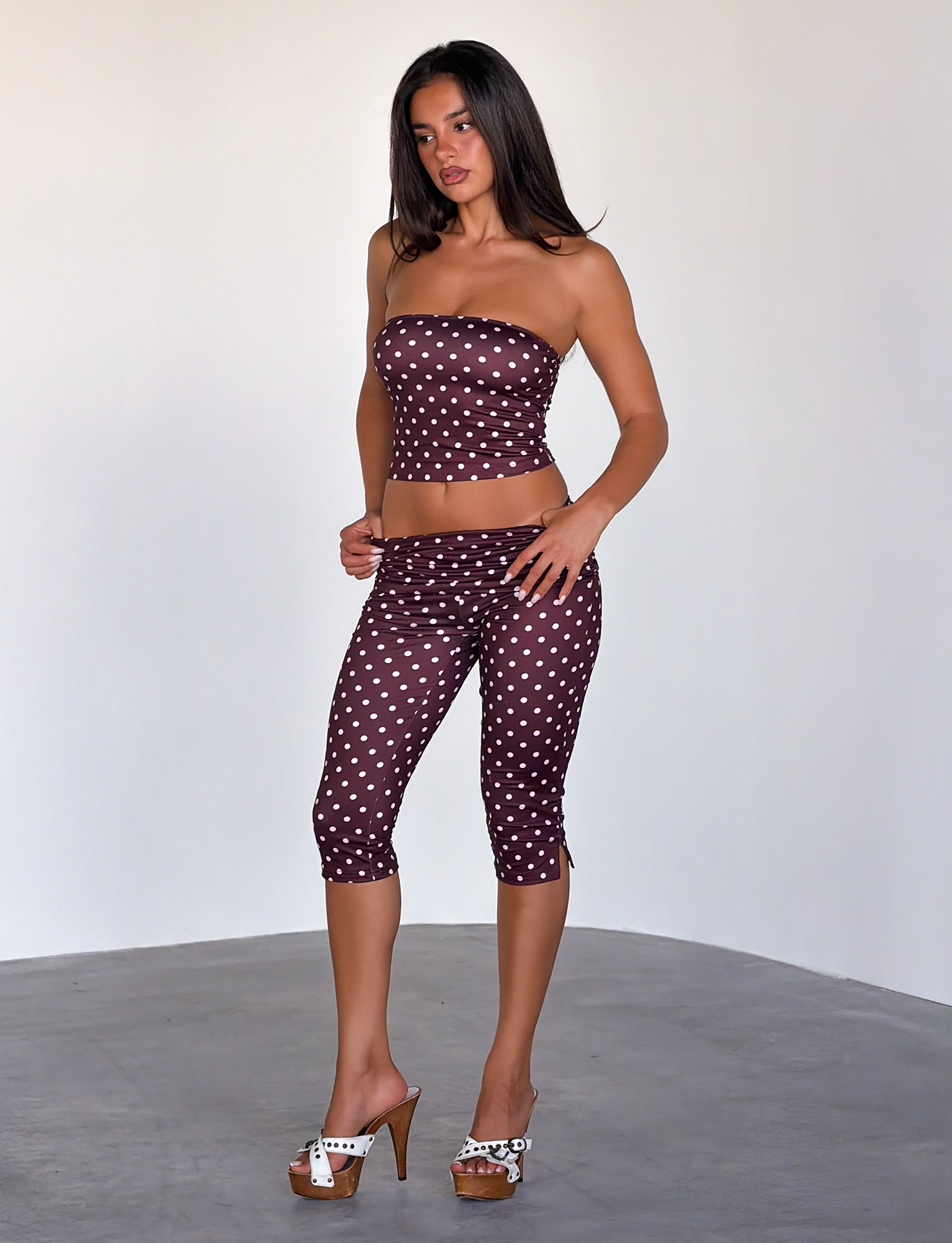 A woman with long dark hair wears the TIGERMIST VALI POLKA DOT TOP in chocolate pink dot and maroon capri leggings, posing indoors with one hand on her hip and high-heeled sandals.