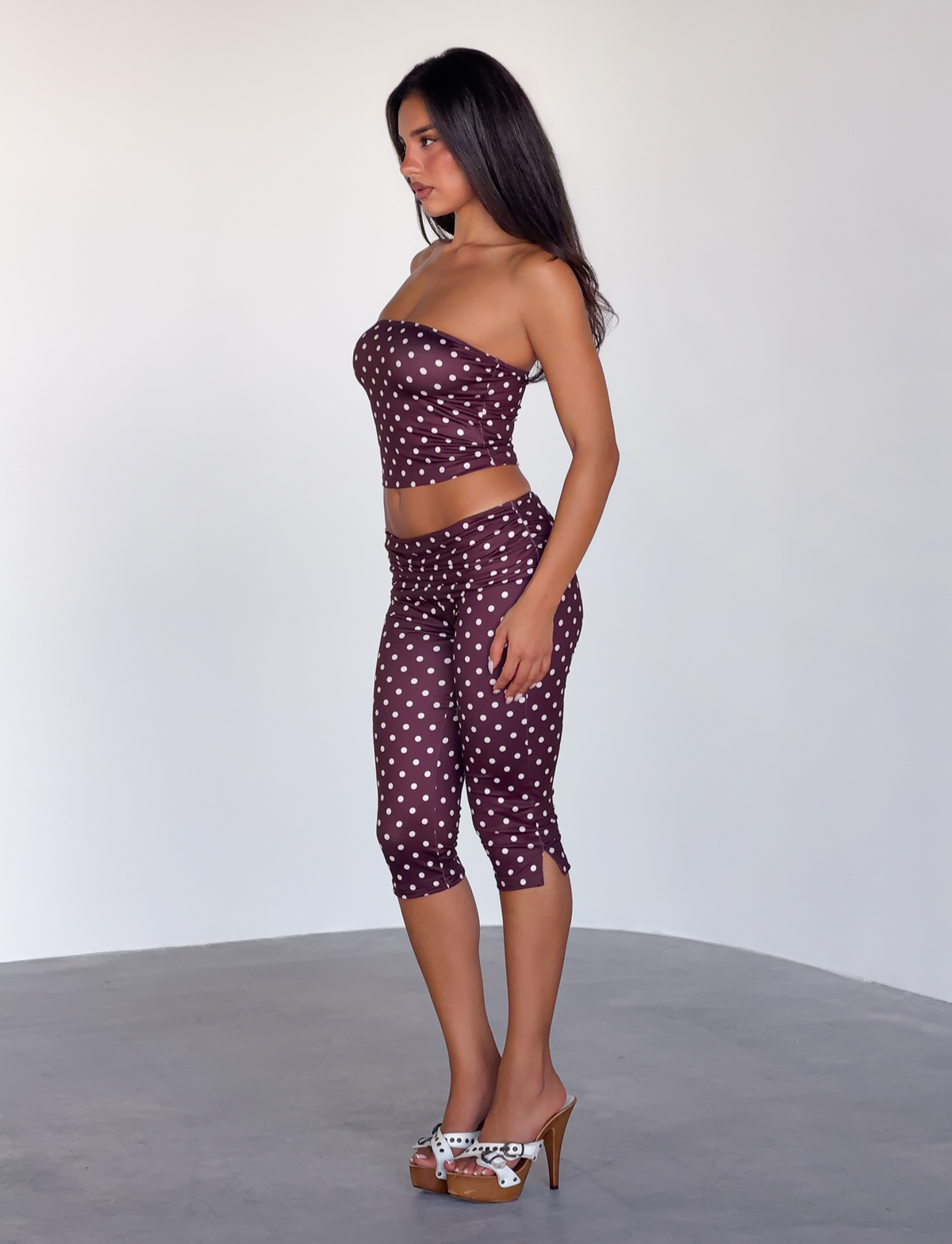 A woman stands in profile against a plain background, wearing the TIGERMIST VALI POLKA DOT TOP in chocolate pink dot with matching capri pants, paired with high-heeled sandals.