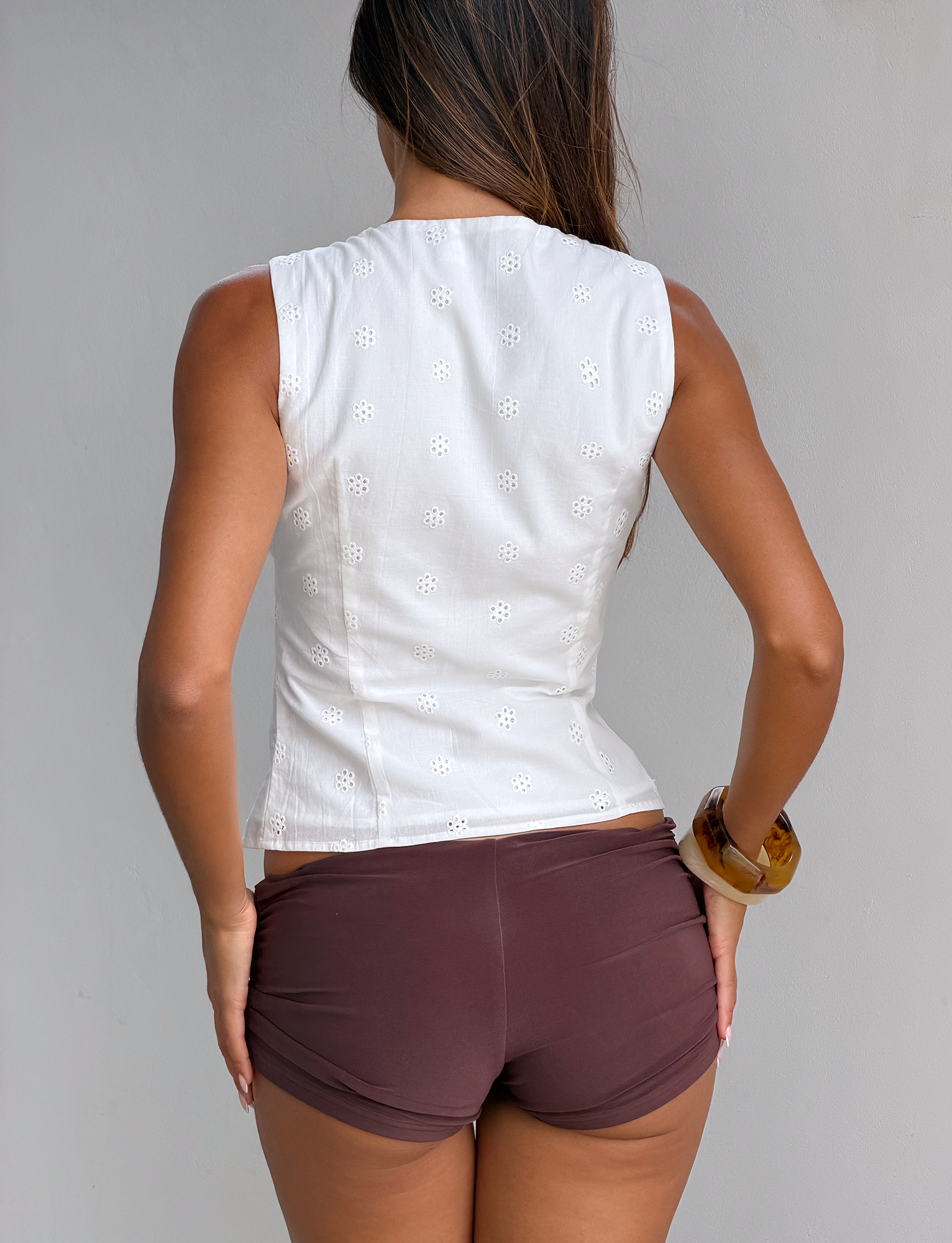 A woman with long brown hair faces away, wearing the TIGERMIST CAELYNN TOP in white featuring subtle eyelet trim and brown shorts, accessorized with a brown bangle on her right wrist, set against a plain light background.