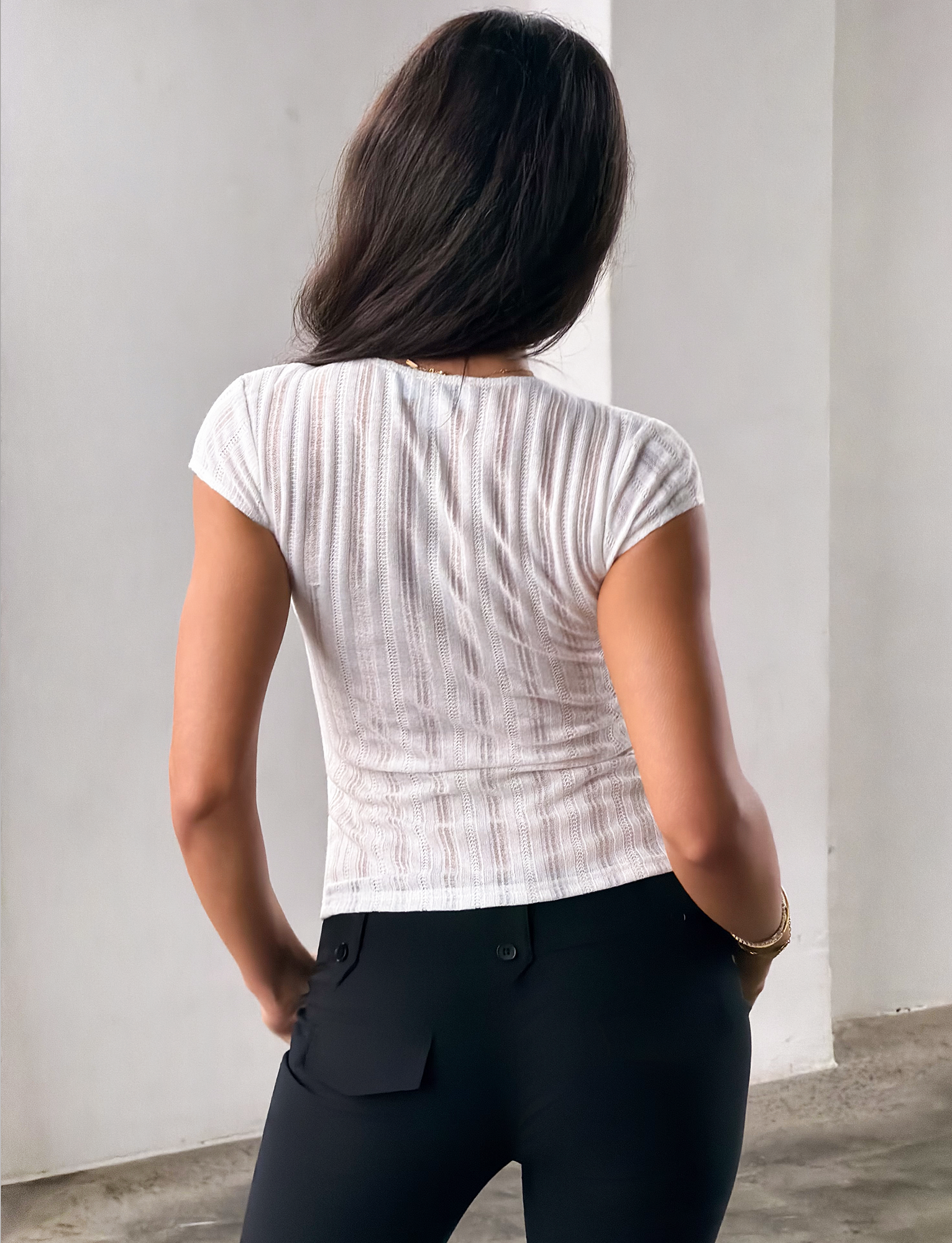 A woman with long brown hair stands indoors, facing away. She is wearing the TIGERMIST LONA TOP in white—a textured V neck top with short sleeves—paired with black pants. The setting features a light-colored wall and a concrete floor.