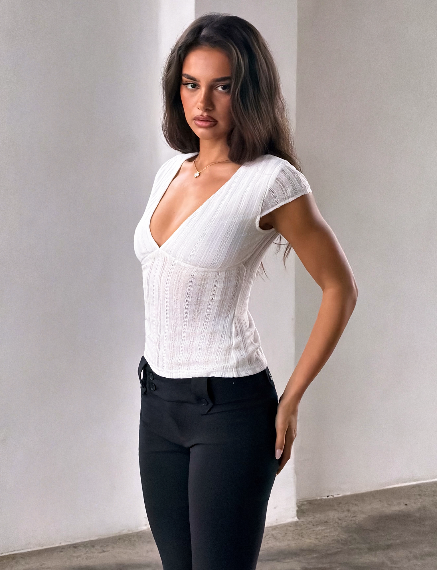 A woman with long brown hair wears the TIGERMIST LONA TOP in white and black pants, standing indoors against a plain light-colored wall, looking at the camera with a neutral expression.