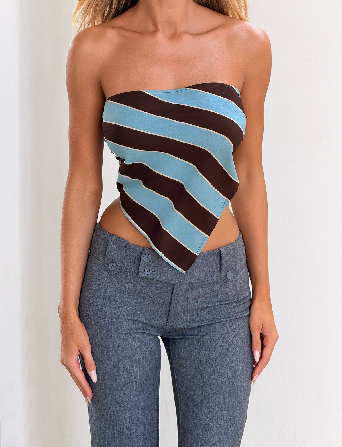 A woman models the TIGERMIST LENIN SCARF TOP - BLUE AND BROWN STRIPE with high-waisted gray pants against a plain white background. Only her torso is visible; her head and lower legs are out of frame.