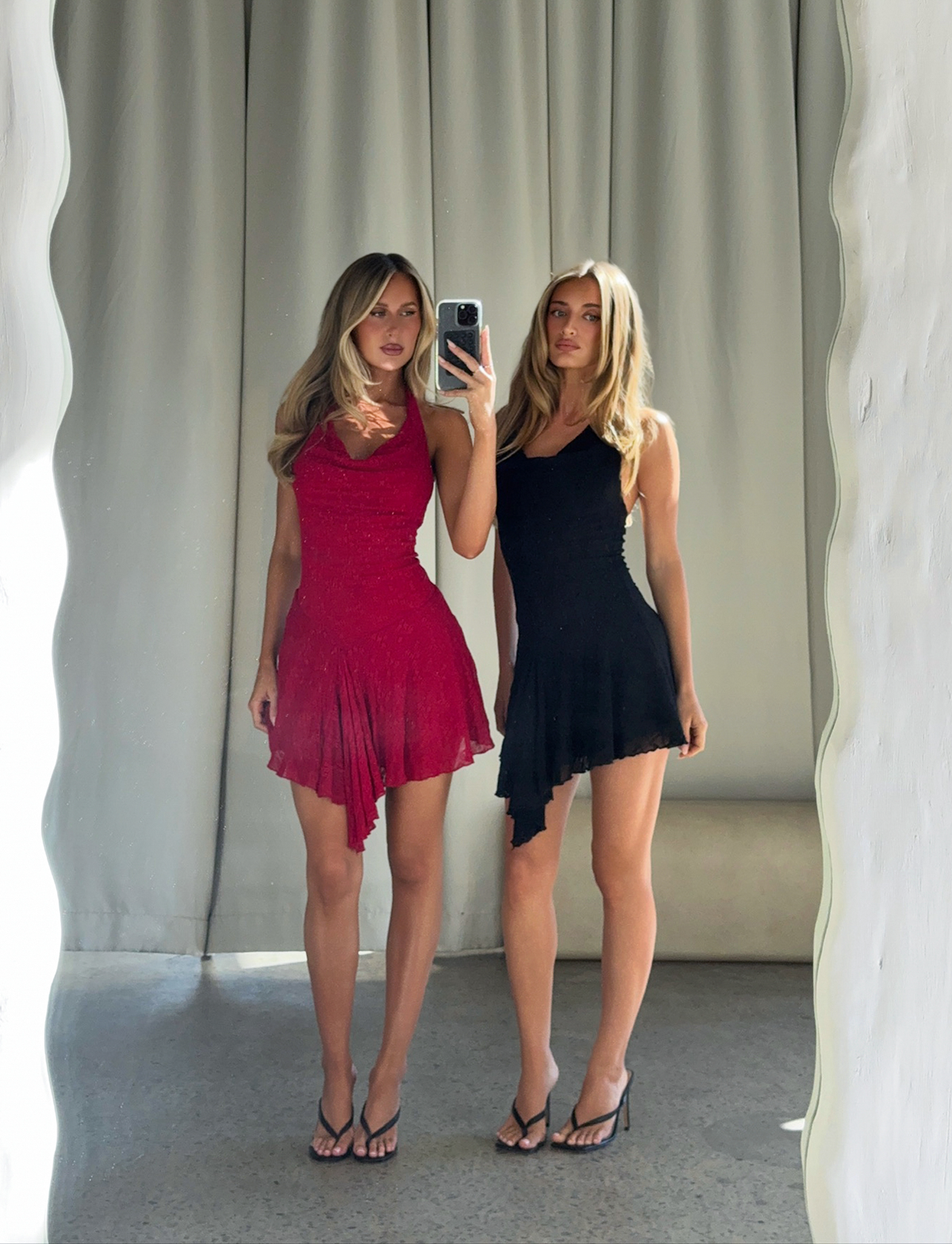 Two women stand by a wavy-edged mirror, one in the TIGERMIST FIORELLA DRESS - RED LACE, the other in a classic black dress. Both have long hair, wear stylish sandals, and are framed by neutral curtains.