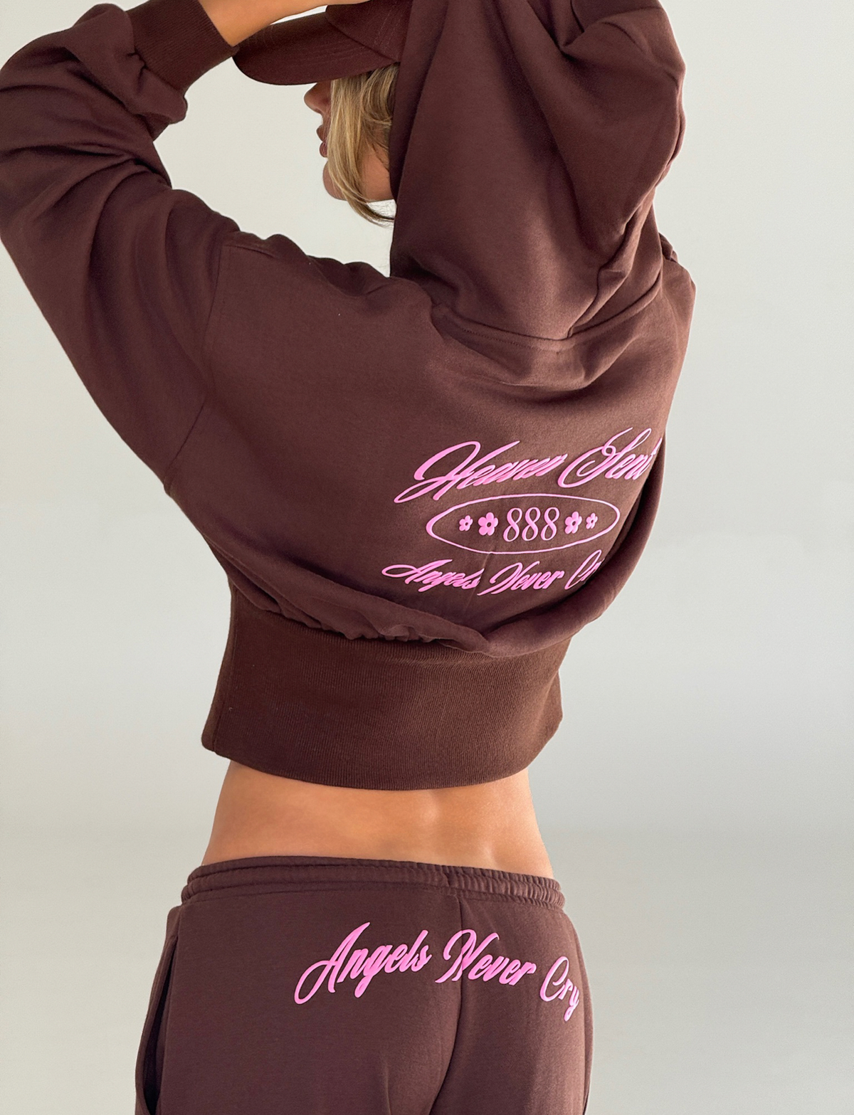 Wearing the TIGERMIST MERRYN HOODIE in Chocolate, featuring pink puff print "Heaven Sent 888" and "Angels Never Cry," a person stands with one arm raised, partly covering their head.