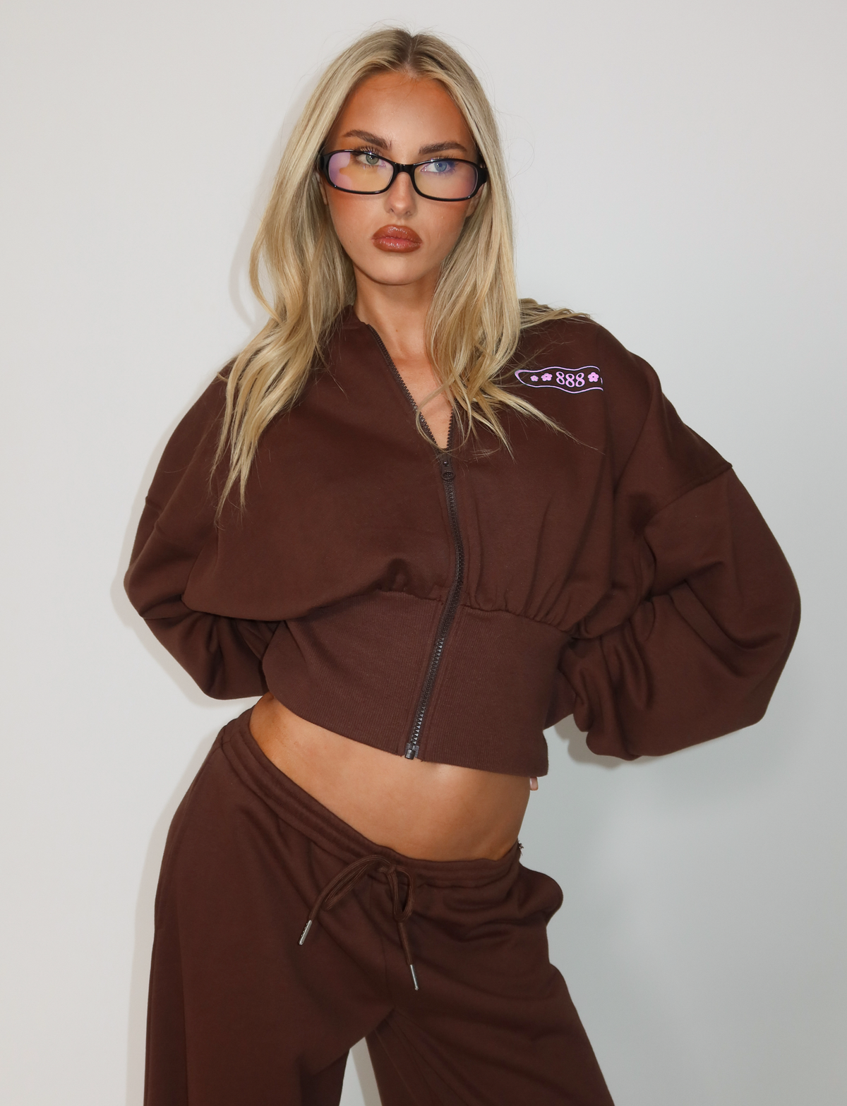 A woman with long blonde hair poses against a light background, wearing dark glasses and the TIGERMIST MERRYN HOODIE - CHOCOLATE with matching pants. The hoodie features a puff print emblem on the upper left side.