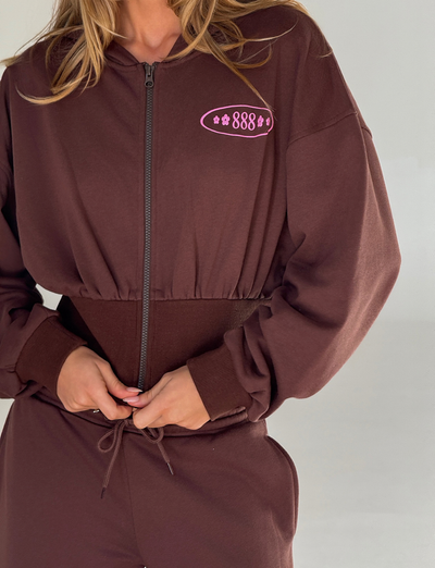 A person wears the TIGERMIST MERRYN HOODIE - CHOCOLATE with matching pants. The lightweight zip-up hoodie displays a pink “888” puff print and stars on the left chest as they adjust the waistband against a plain, light background.
