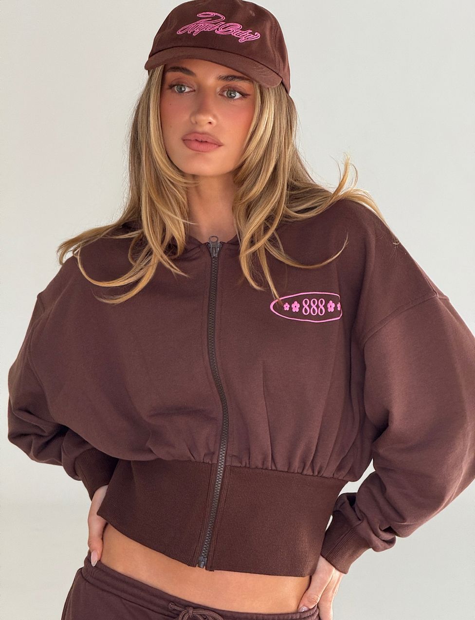 A woman poses with one hand on her hip, wearing the TIGERMIST MERRYN HOODIE in chocolate, a brown lightweight zip-up with pink embroidered text, against a plain light background.