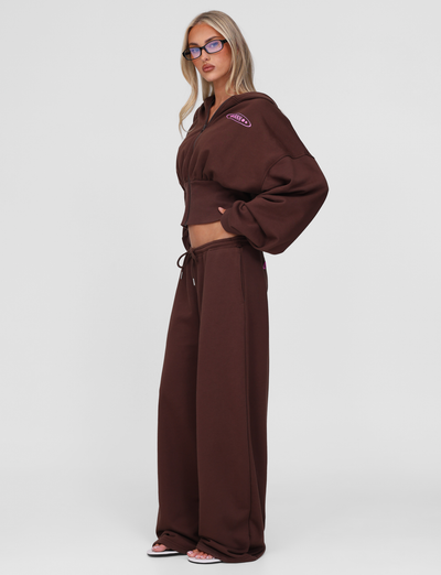 A woman with long blonde hair and glasses stands sideways, barefoot against a white background, wearing the TIGERMIST MERRYN HOODIE - CHOCOLATE and matching wide-leg sweatpants.