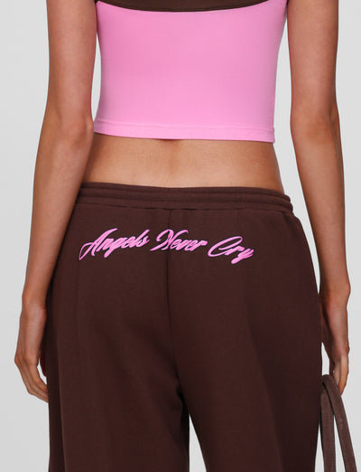 A person wears TIGERMIST’s MERRYN TRACK PANT in chocolate, featuring a pink "Angels Never Cry" print on the back. The image is cropped to show only their back and waist.