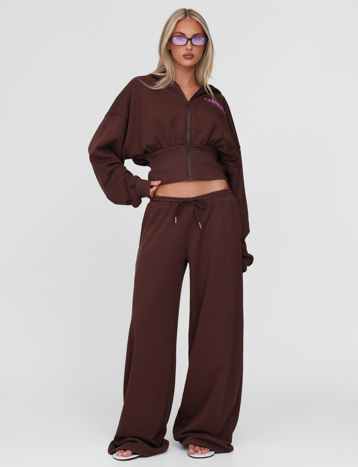 A woman with long blonde hair and purple glasses stands against a white background wearing TIGERMIST’s MERRYN TRACK PANT - CHOCOLATE, paired with white sandals. Her hands on her waist embody the Angels Never Cry spirit.