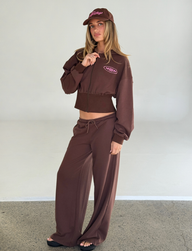 A woman with long hair poses indoors against a light background, wearing TIGERMIST’s MERRYN TRACK PANT - CHOCOLATE with a matching brown zip-up hoodie, brown cap, and black flip-flops. She has a neutral expression.