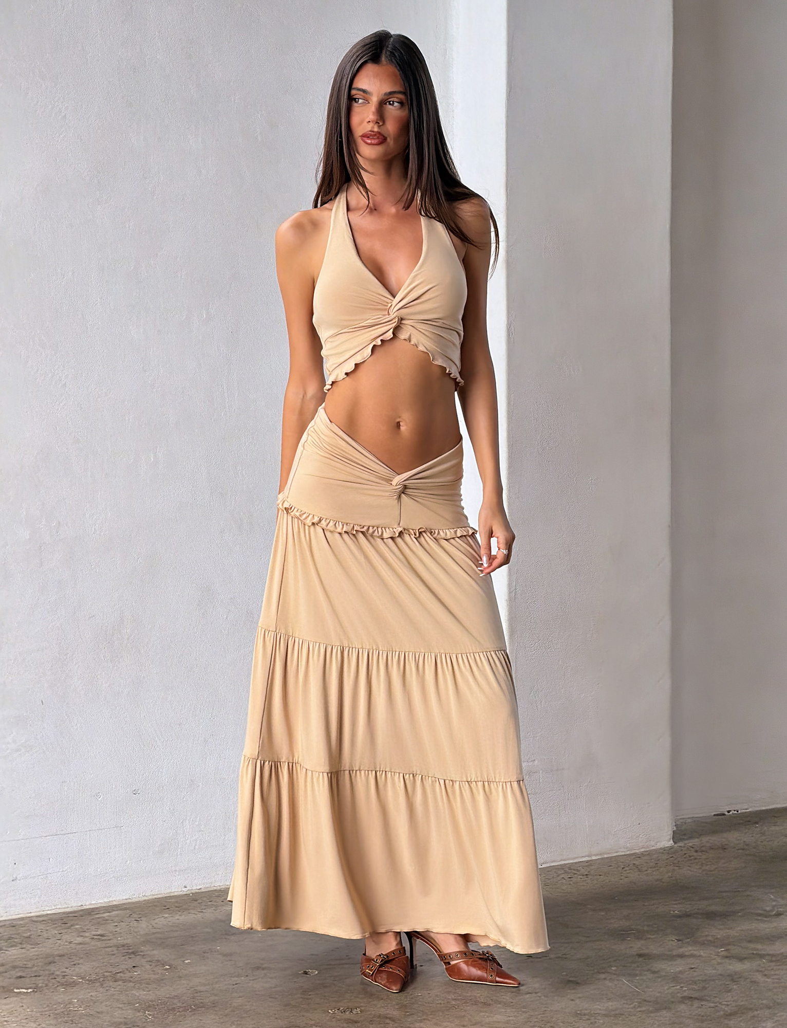 A woman stands indoors wearing the TIGERMIST KATYA MAXI SKIRT in tan, styled with a beige halter crop top. She has long straight hair, brown flat pointed shoes, and the background is minimal.