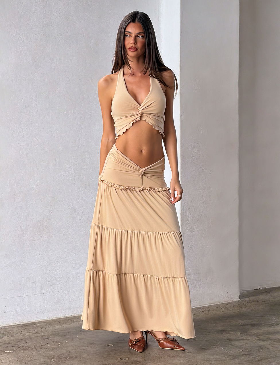 A woman stands indoors wearing the TIGERMIST KATYA MAXI SKIRT in tan, styled with a beige halter crop top. She has long straight hair, brown flat pointed shoes, and the background is minimal.