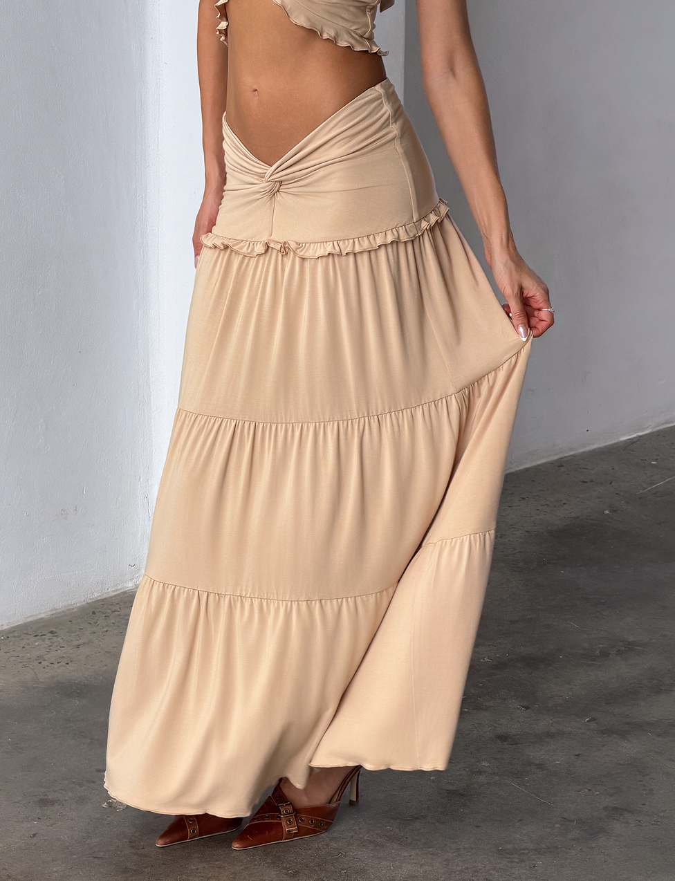 A woman stands indoors on a concrete floor, holding out her KATYA MAXI SKIRT - TAN by TIGERMIST. The beige, tiered maxi skirt features a wide waistband; her head and most of her upper body are not visible in the image.