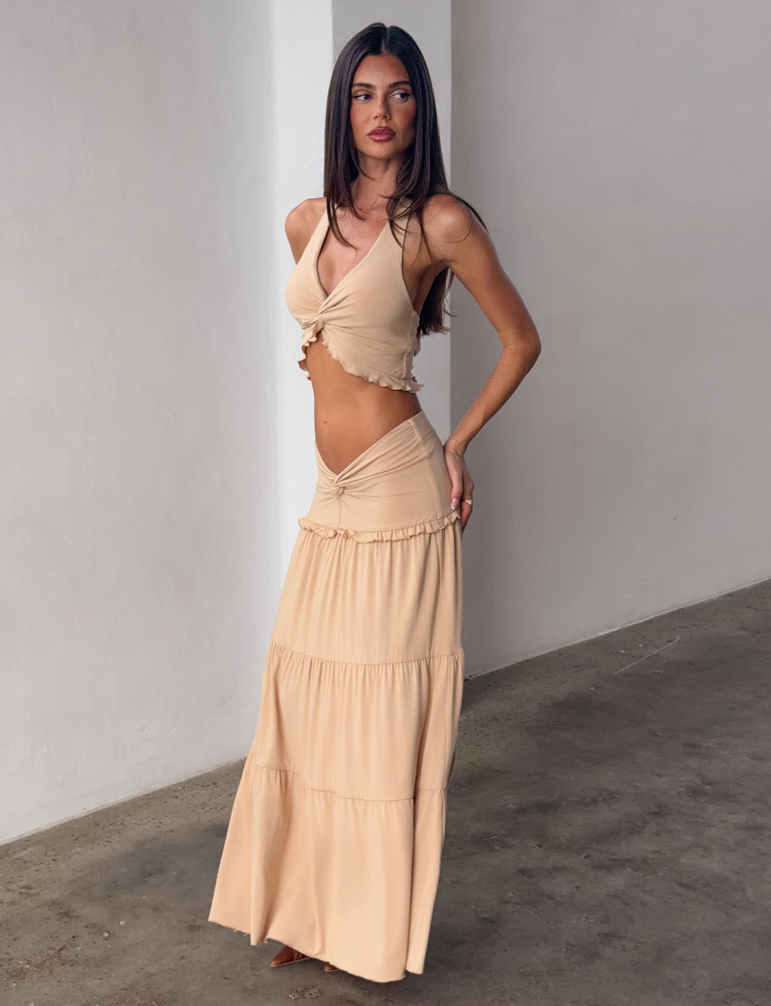 A woman with long brown hair wears the TIGERMIST KATYA MAXI SKIRT in tan, paired with a beige sleeveless crop top, standing barefoot on a concrete floor against a plain white wall.