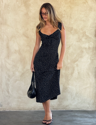 A woman with long hair and glasses wears the LOTTIE RUFFLE MIDI DRESS - BLACK POLKA by TOP ONE LADYS FASHION PTY LTD, styled with heeled sandals and a black handbag, standing against a beige textured wall.
