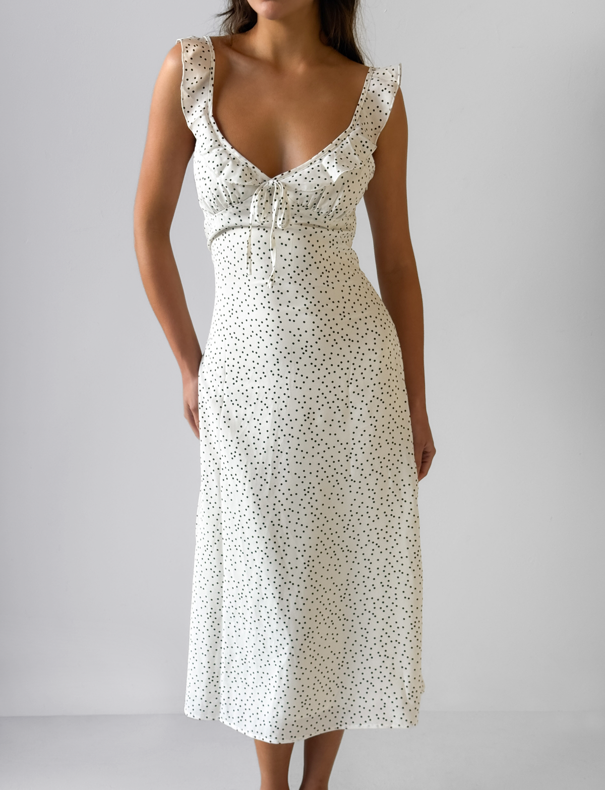 A woman wears the CL - LOTTIE RUFFLE MIDI DRESS by Tiger Mist, a sleeveless white dress with black polka dots, ruffled V-neckline, chest tie detail, fitted waist, and gently flowing midi skirt against a plain light gray background.