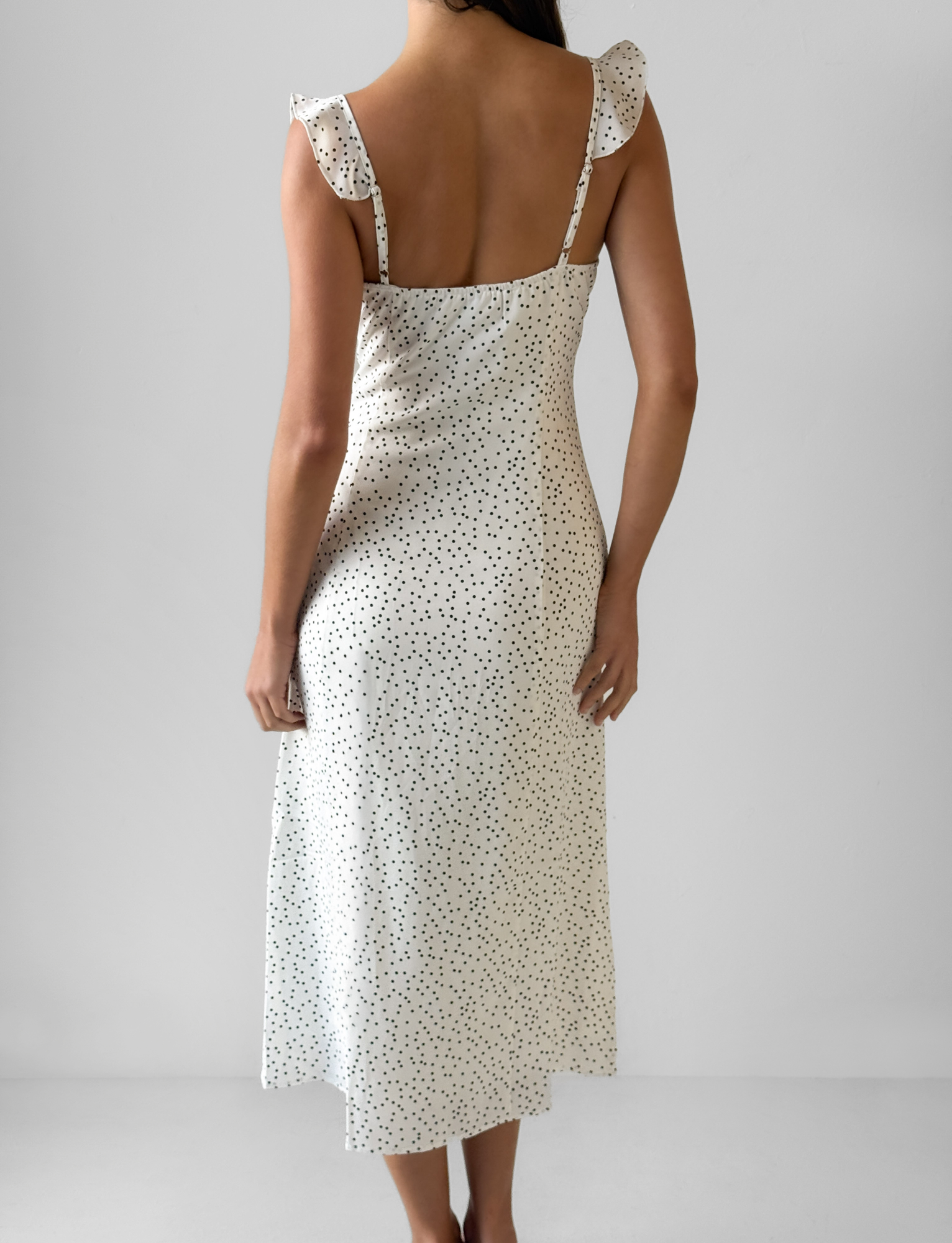 A woman faces away in the LOTTIE RUFFLE MIDI DRESS - WHITE POLKA by TOP ONE LADYS FASHION PTY LTD, featuring ruffled shoulder straps, set against a plain light gray background.