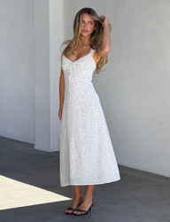 A woman stands indoors against a light gray wall, wearing the LOTTIE RUFFLE MIDI DRESS - WHITE POLKA by TOP ONE LADYS FASHION PTY LTD and black sandals, looking to the side with one hand touching her hair.