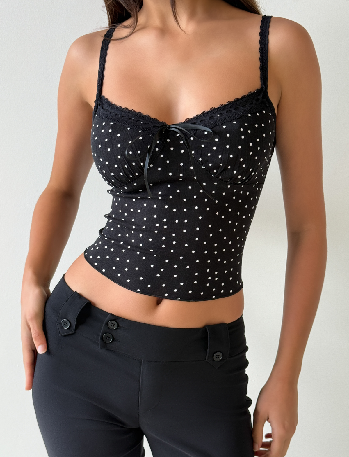 A woman models the Tiger Mist CL - BAILEE CAMI TOP, a black, sleeveless polka dot piece with lace trim and bow detail, paired with black high-waisted pants. She stands against a light background, face not visible.