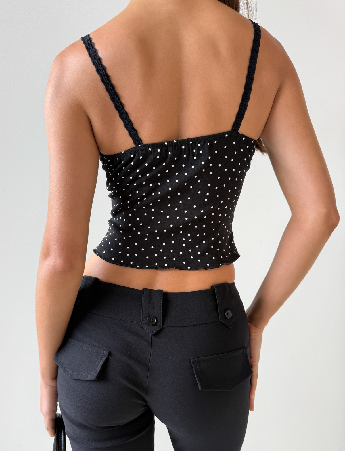 A person is shown from the back wearing the BAILEE CAMI TOP - BLACK POLKA by TOP ONE LADYS FASHION PTY LTD, featuring white polka dots and lace trim, paired with black pants against a plain light background.