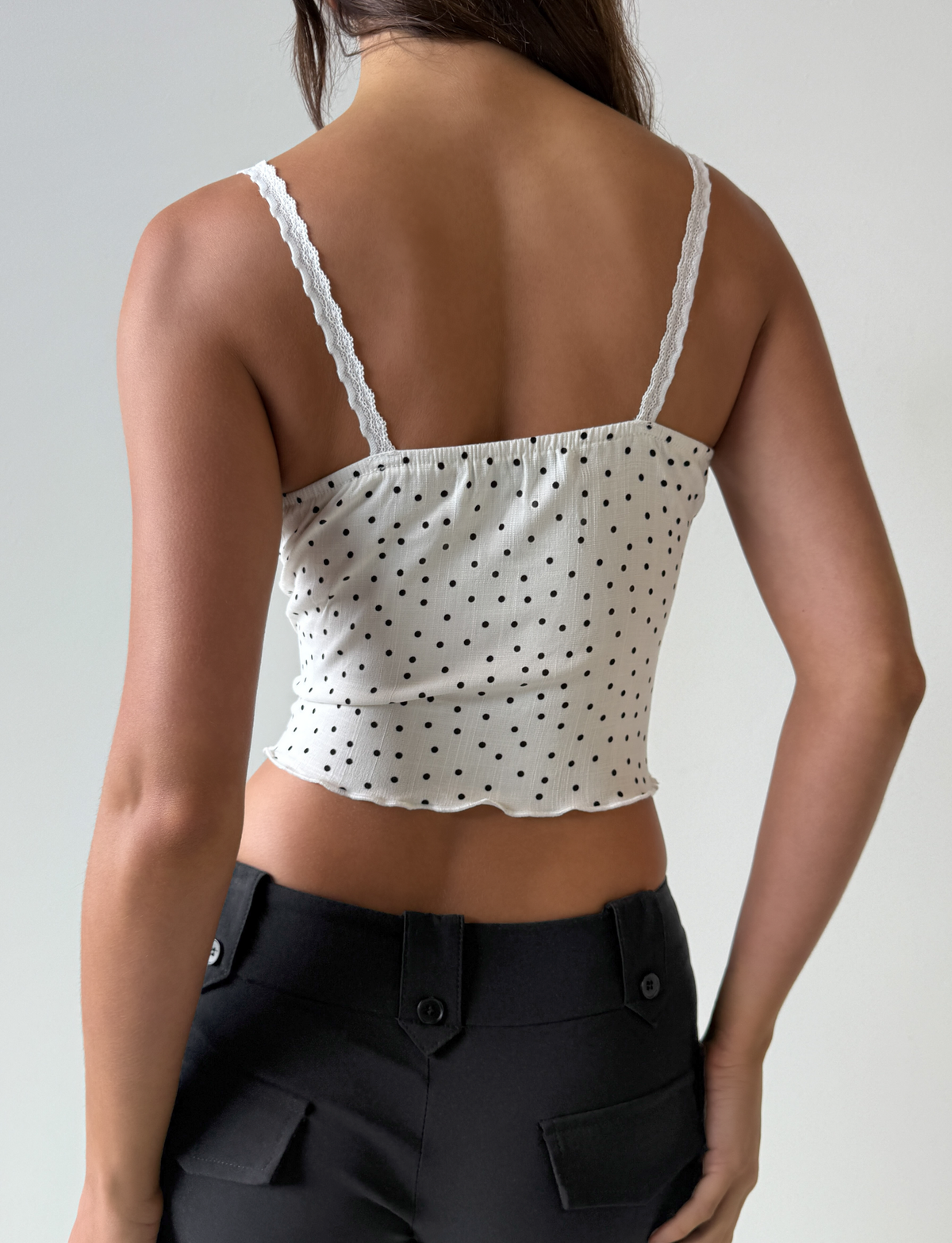 A woman wears the BAILEE CAMI TOP - WHITE POLKA by TOP ONE LADYS FASHION PTY LTD, featuring delicate lace trim, paired with black pants.
